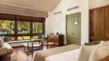 A room with a bed, a sofa, a dining table, and chairs. There is a window with a view of greenery outside.