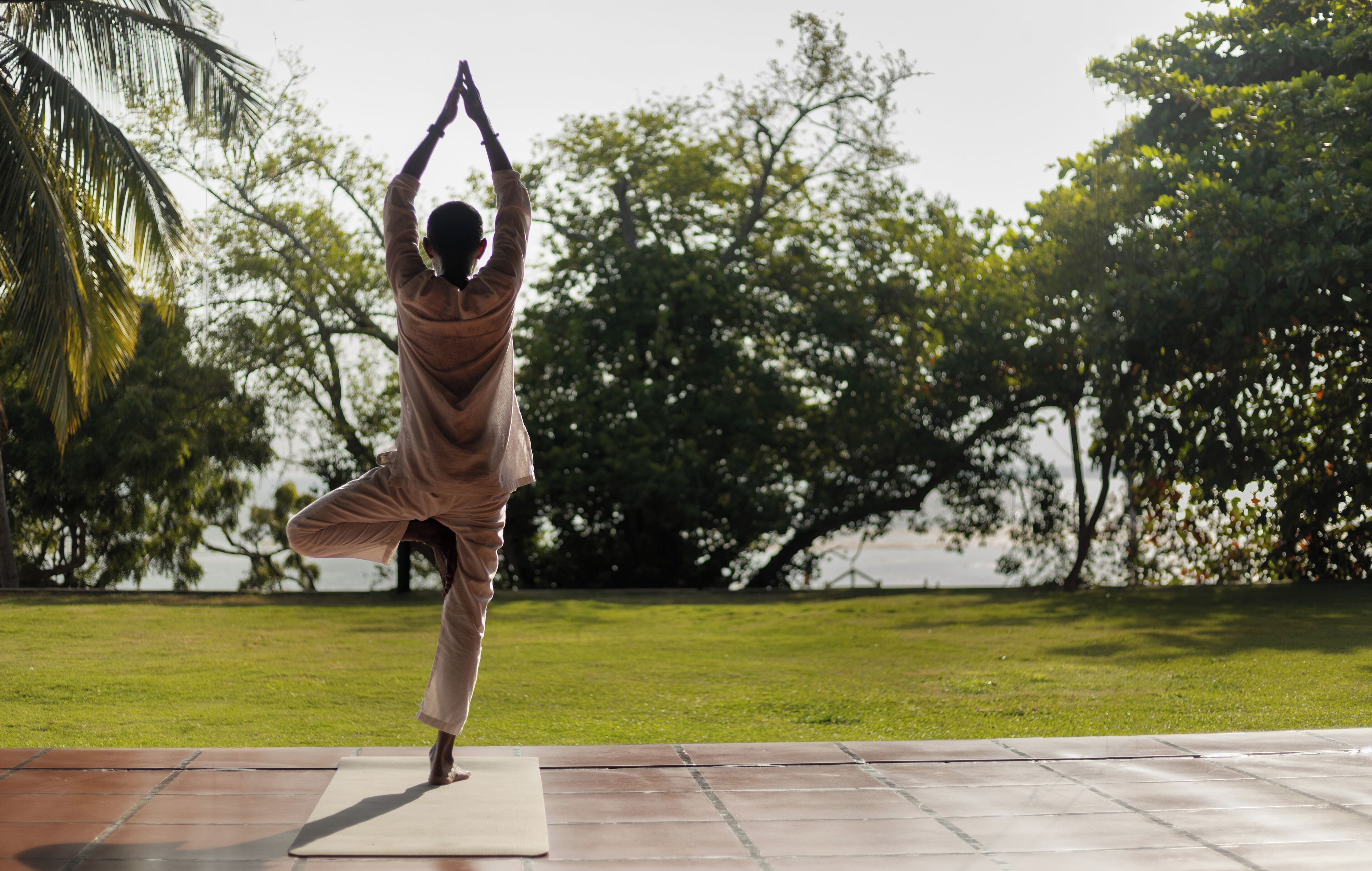A person performing a yoga pose on a mat outdoors with trees and a body of water in the background.