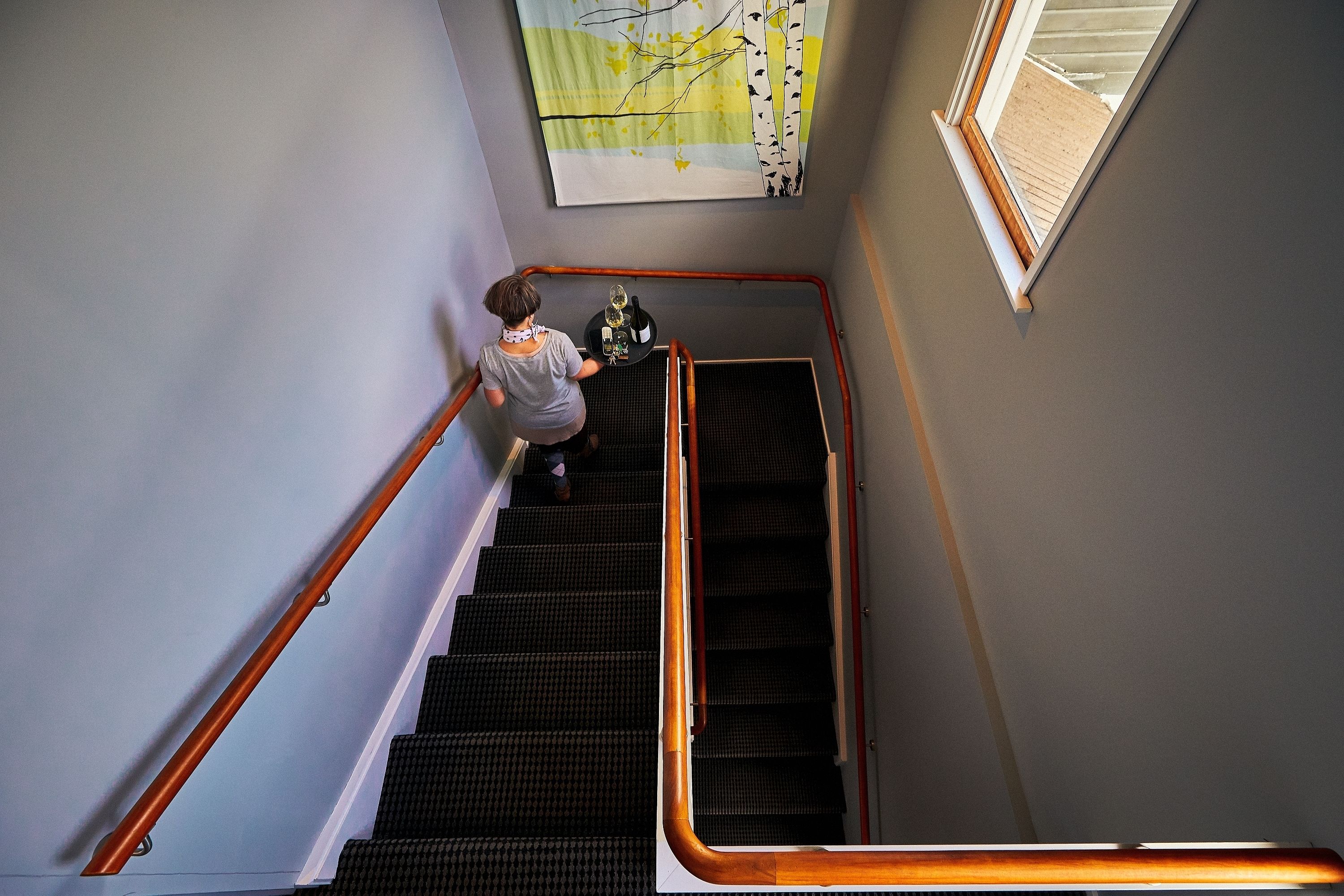 A person descending a staircase with a handrail, a window with a view of a wooden deck, and a wall-mounted artwork.