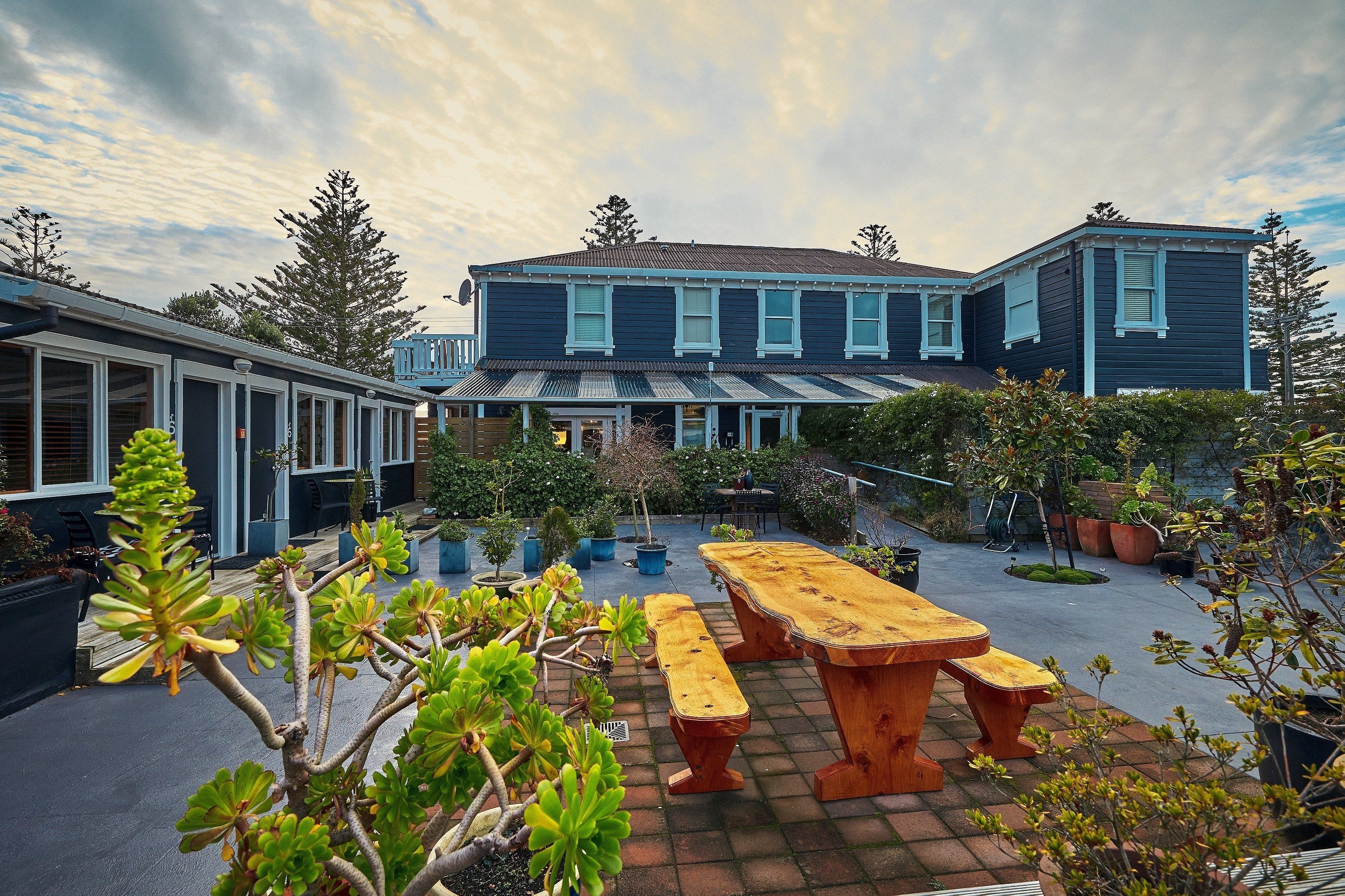 A courtyard with a wooden table and benches, surrounded by potted plants and trees.