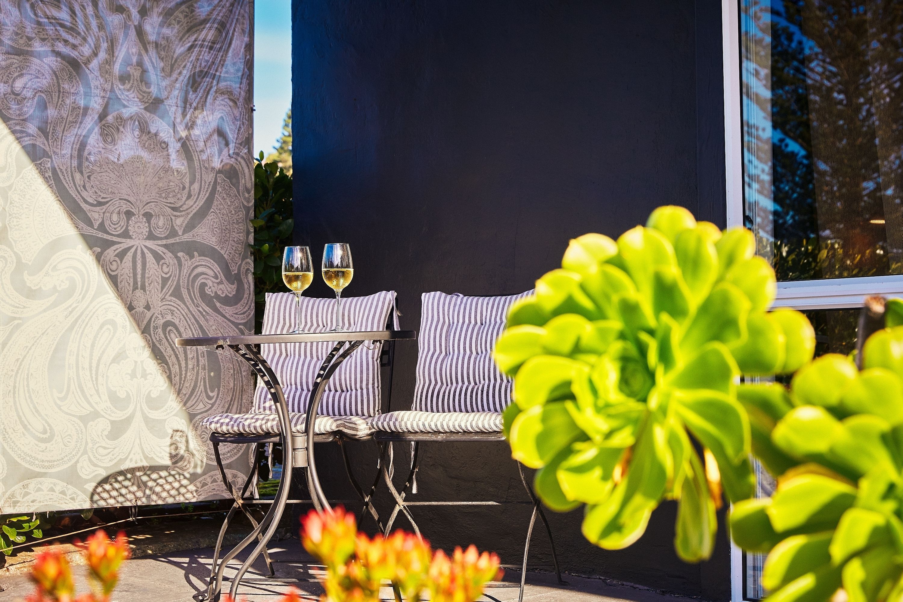 An outdoor seating area with two chairs and a table, two glasses of white wine, and a patterned wall.