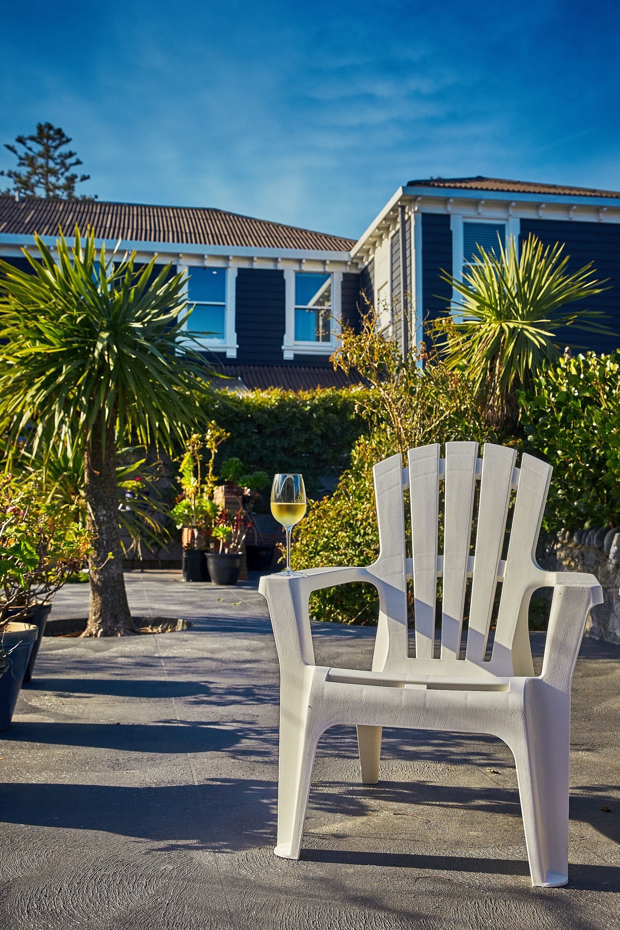 A white plastic chair with a glass of white wine on it, placed on a concrete patio with potted plants and a building in the background.
