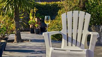 A white plastic chair with a glass of white wine on it, placed on a concrete patio with potted plants and a building in the background.