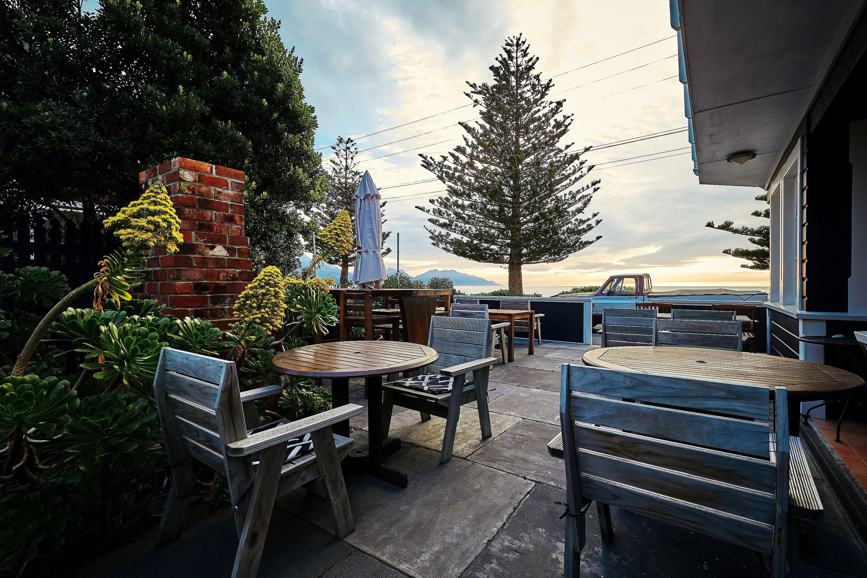 An outdoor patio with wooden tables and chairs, surrounded by greenery and a brick structure.