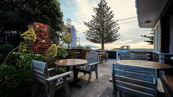 An outdoor patio with wooden tables and chairs, surrounded by greenery and a brick structure.