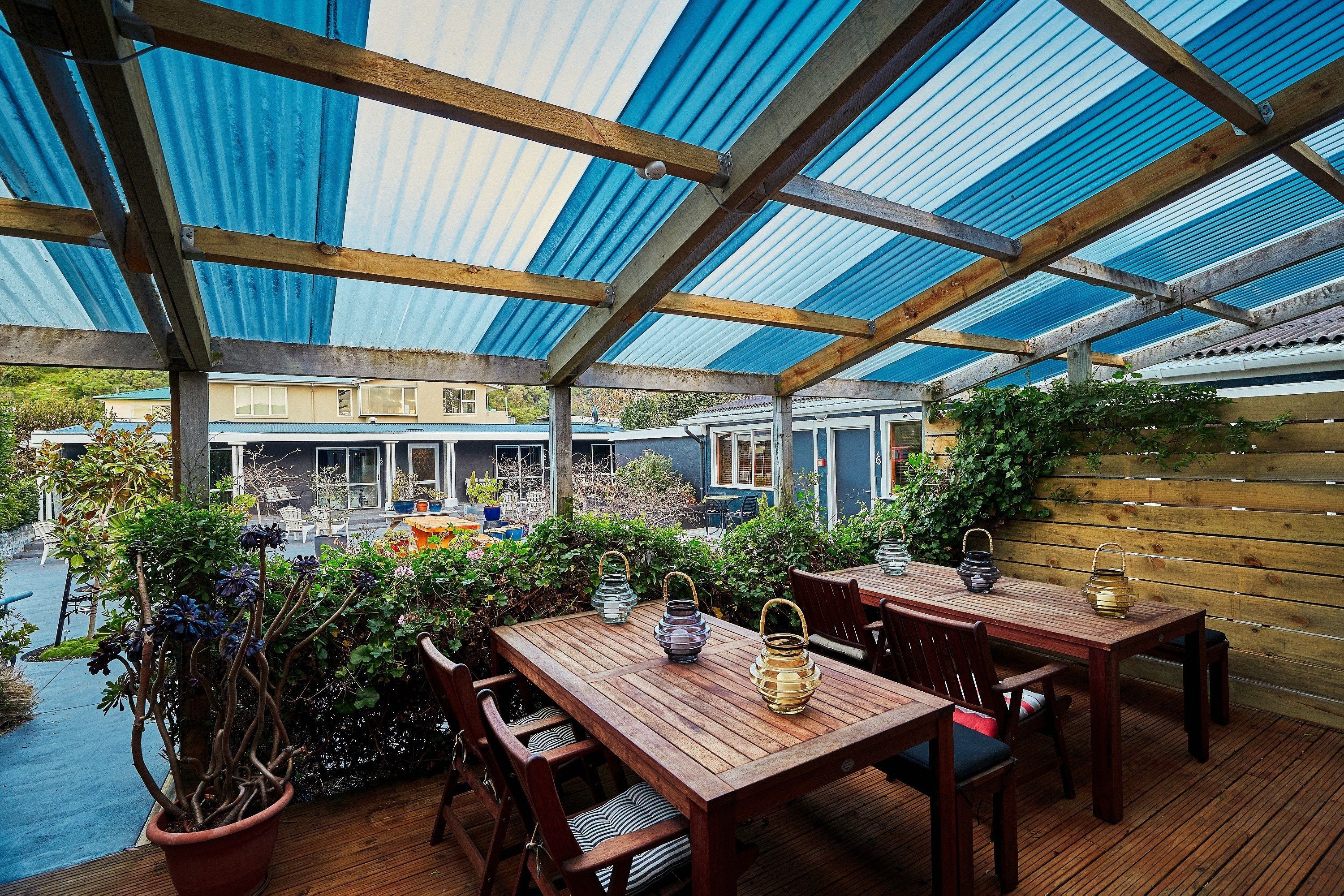 A covered outdoor patio with wooden tables and chairs, potted plants, and a view of a residential area.