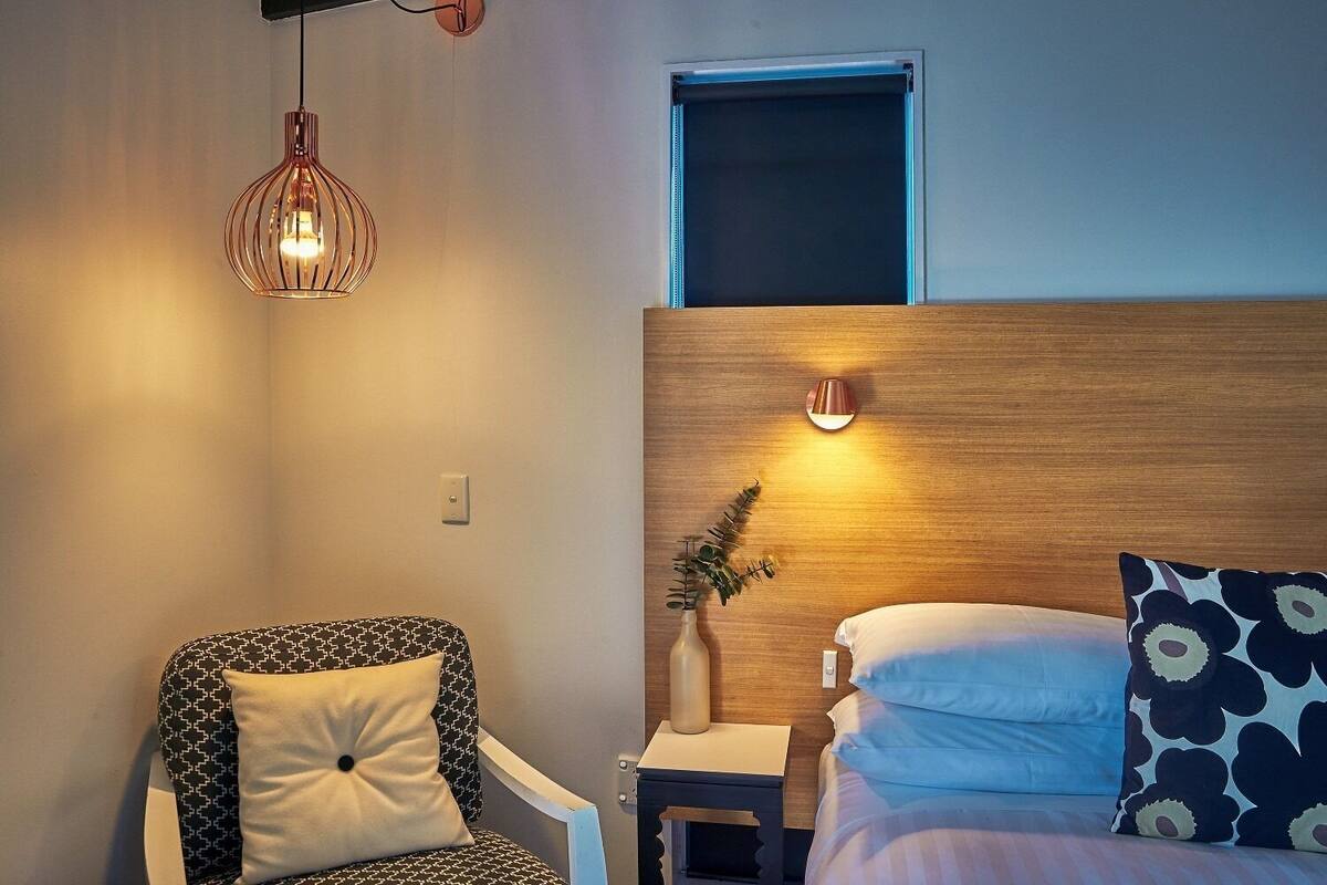 Una habitación de hotel con cama, una silla, una mesita de noche, una lámpara de pared y un ventanal.