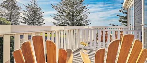 A wooden deck with Adirondack chairs and a glass of wine, overlooking a white fence and tall trees.