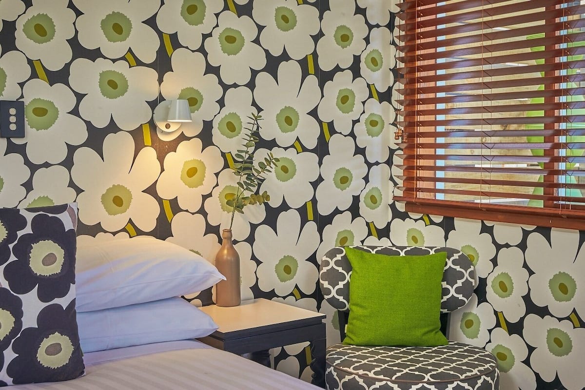 Un dormitorio con papel tapiz floral, una mesita de noche con lámpara, un cojín verde y una ventana con persianas.