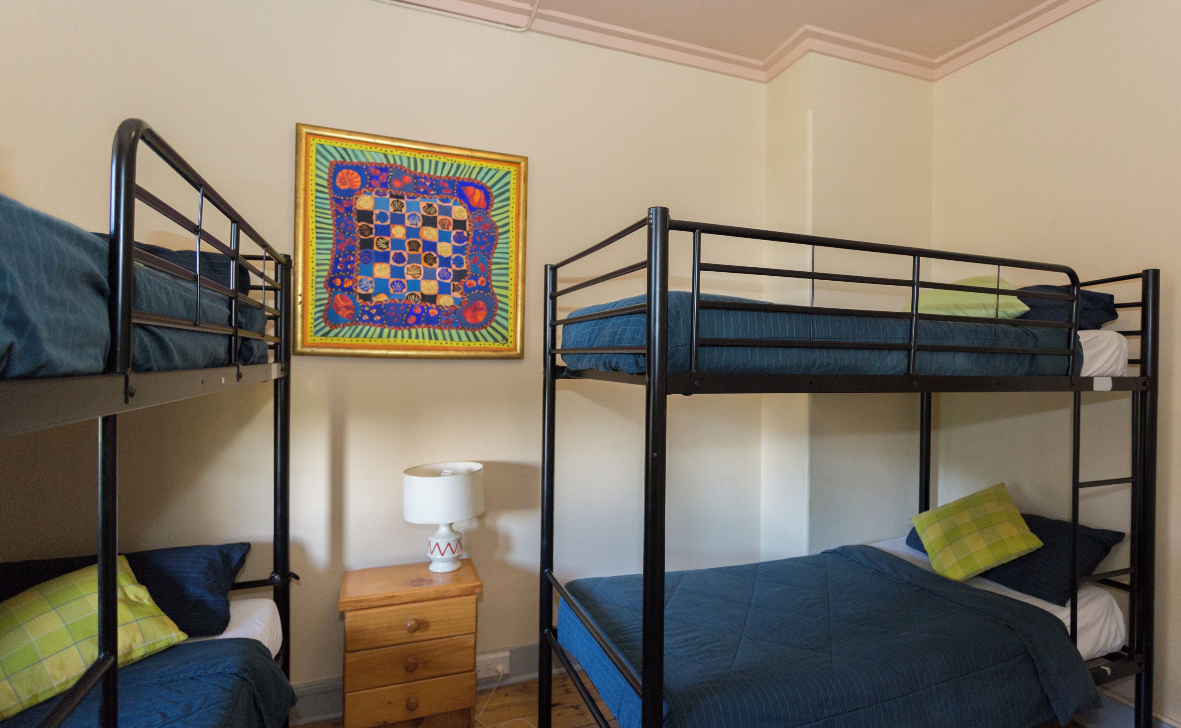 Una habitación con literas, una mesita de noche de madera, una lámpara y un colcha de colores en la pared.