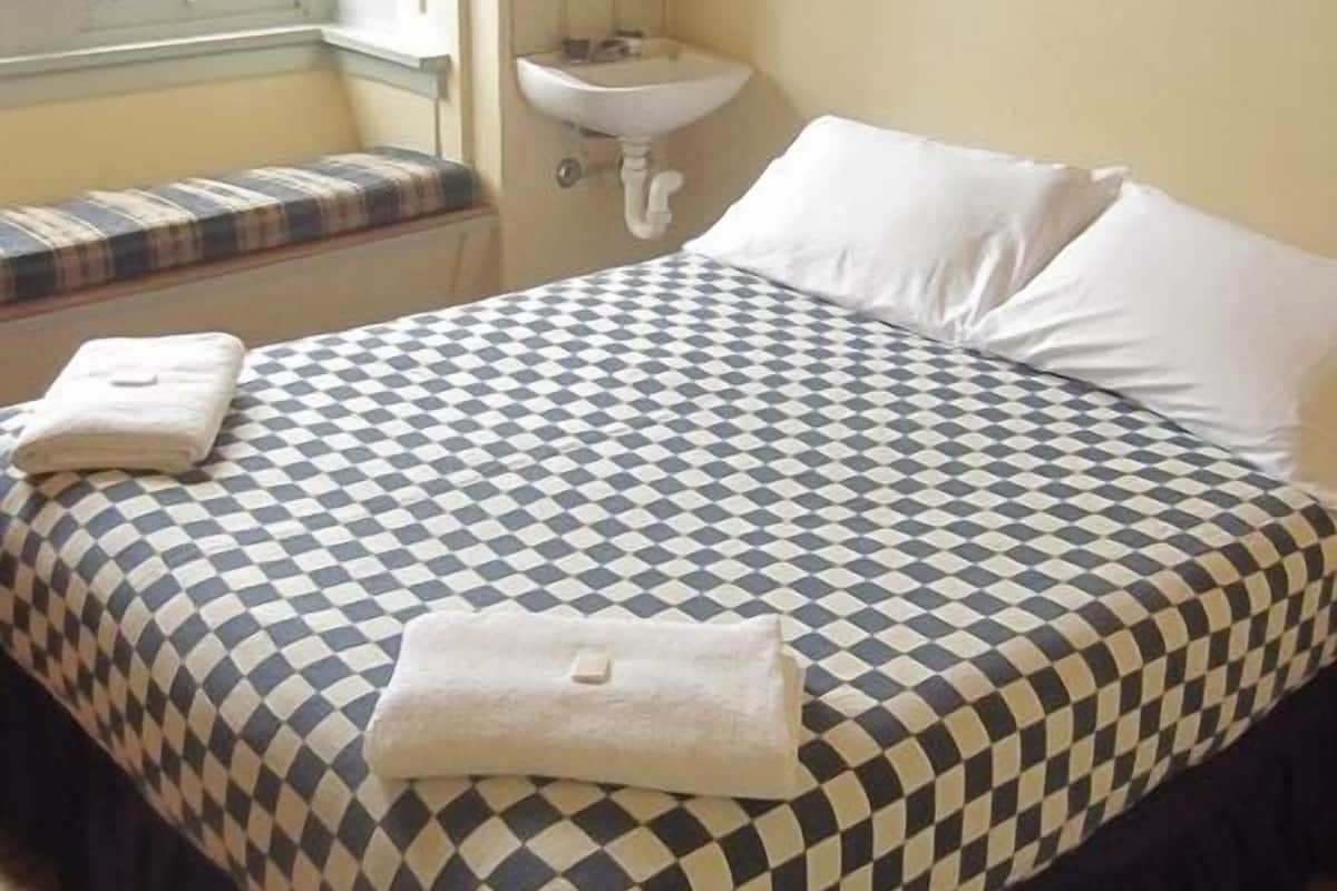 A bedroom with a checkered bedspread, a bedside table with a lamp, a window with a view of the outside, and a painting on the wall.