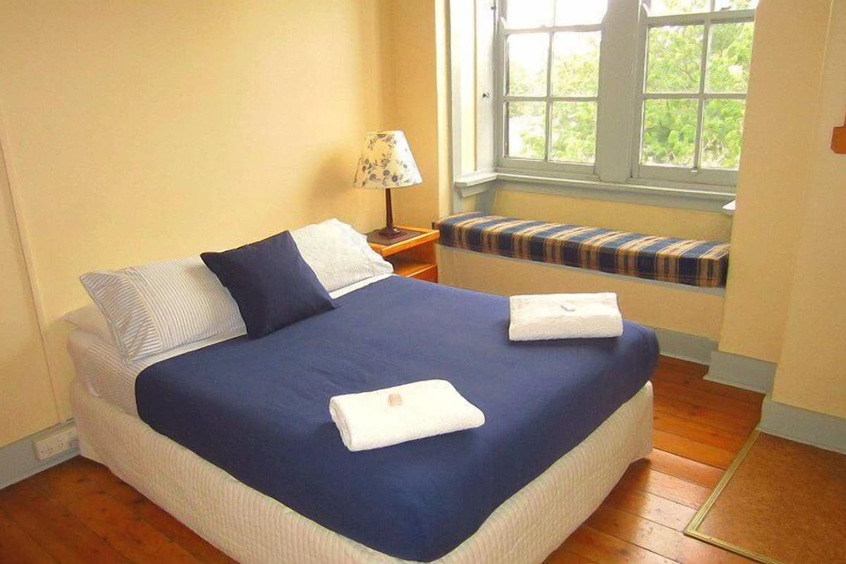 A neatly made bed with a blue bedspread and white pillows, a bedside lamp, and a bench with a striped cushion by a window.