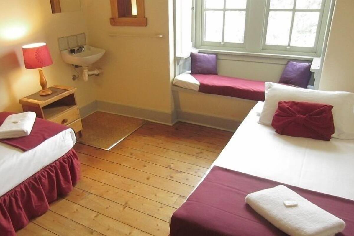 A room with two beds, a nightstand with a lamp, a window with a view, and a small bathroom with a sink.