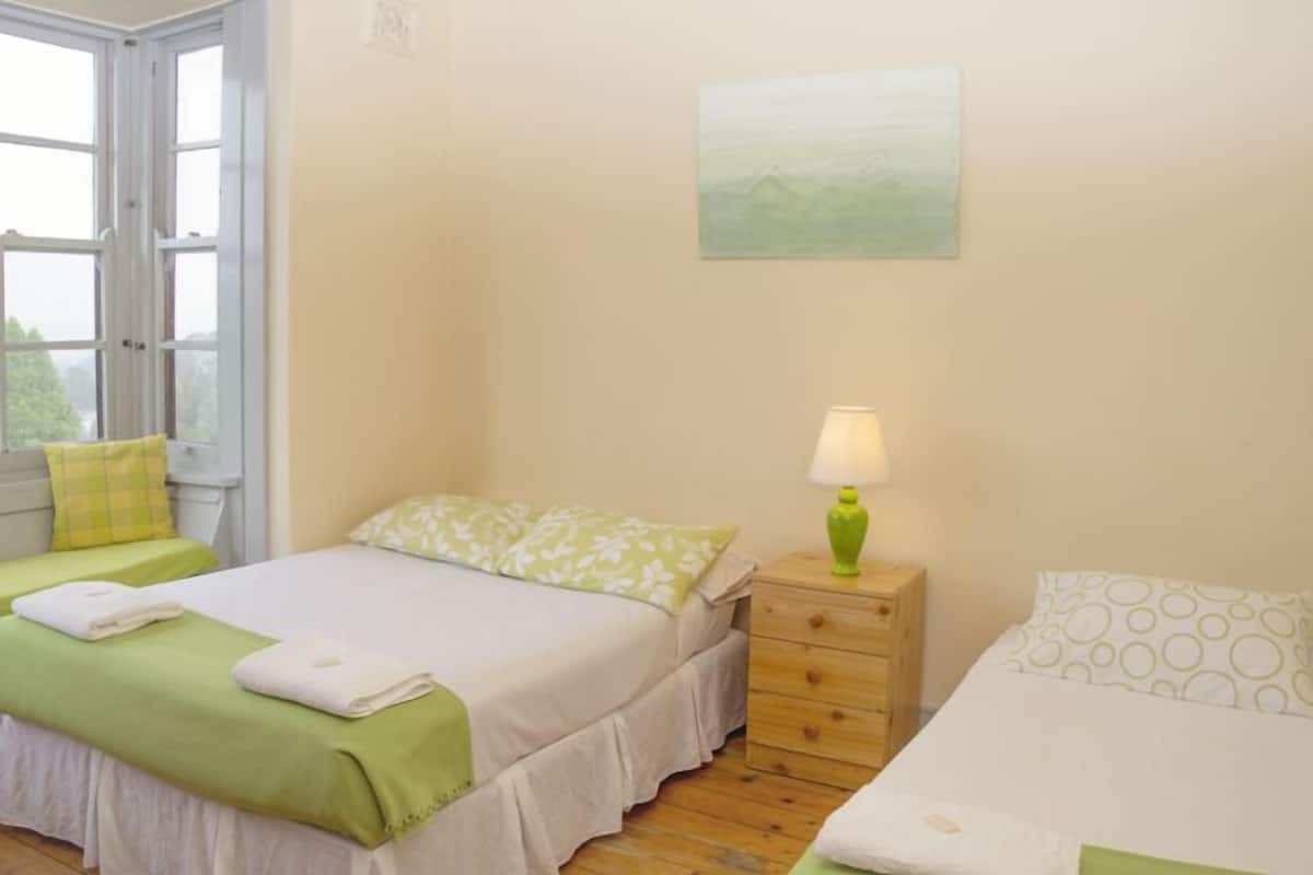A bedroom with two beds, a window, a bedside table with a lamp, and a painting on the wall.