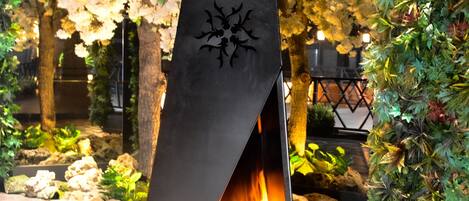 Una chimenea exterior moderna con diseño geométrico, rodeada por un jardín con diversas plantas y un árbol en flor.