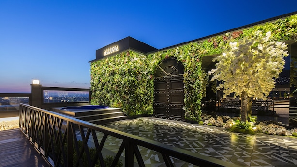 Une terrasse extérieure dotée d’un sol carrelé aux motifs géométriques, d’une plateforme en bois et d’un mur recouvert de verdure et de fleurs blanches.