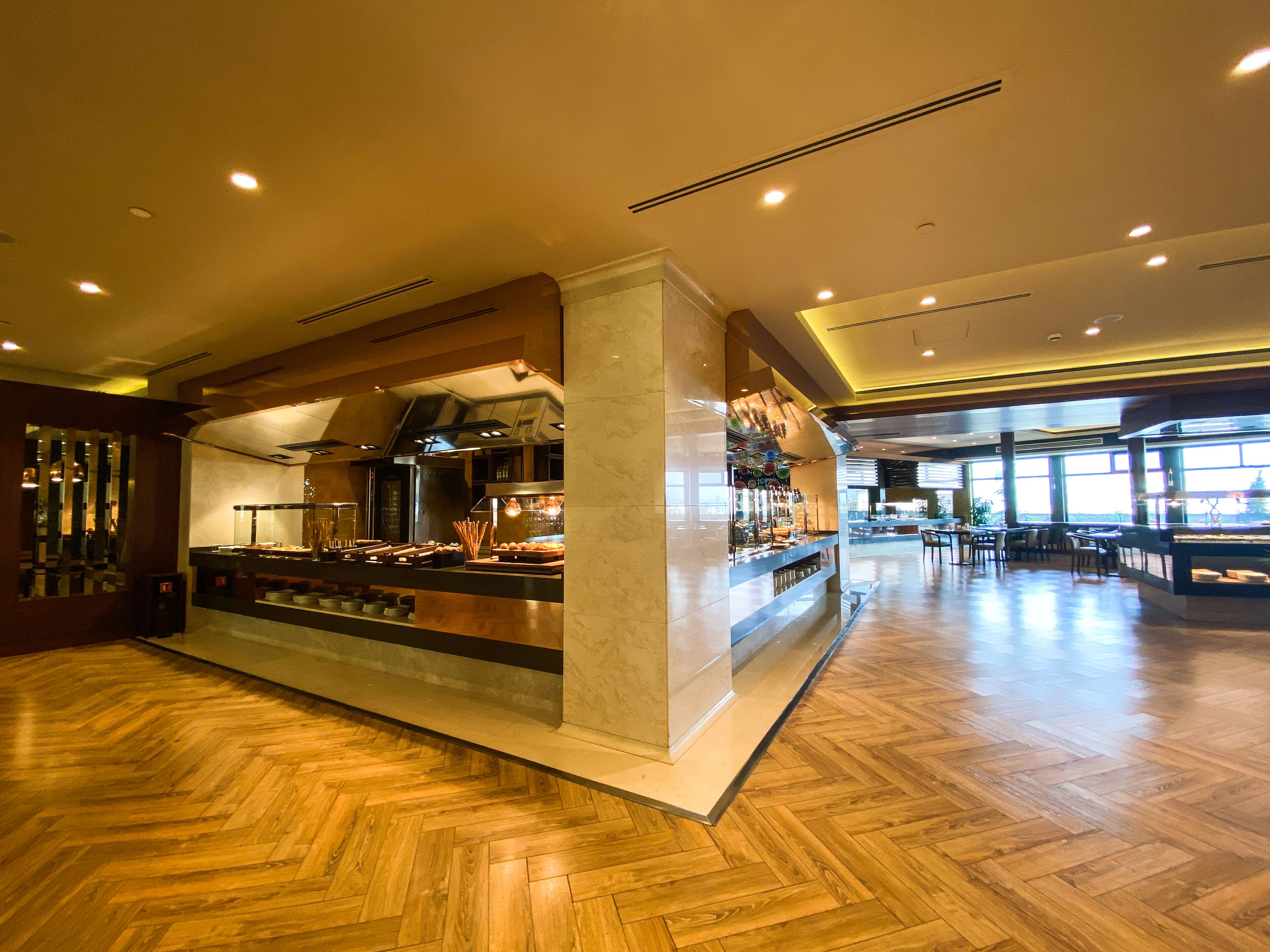 A spacious hotel lobby with a central bar area, wooden flooring, and large windows allowing natural light.