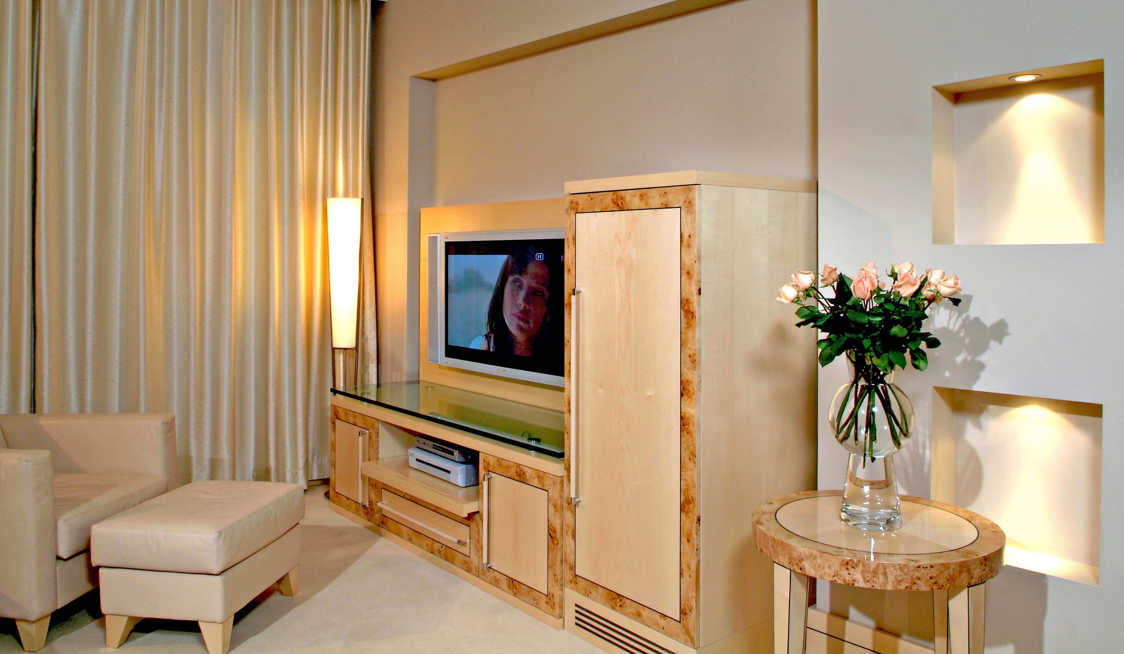 A modern living room with a flat-screen TV, a sofa, a small table with a vase of flowers, and a wall-mounted lamp.
