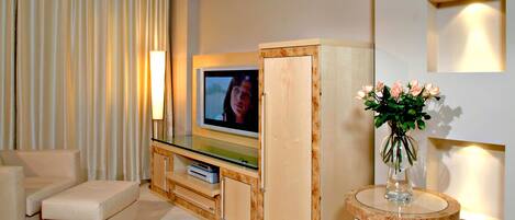 A modern living room with a flat-screen TV, a sofa, a small table with a vase of flowers, and a wall-mounted lamp.