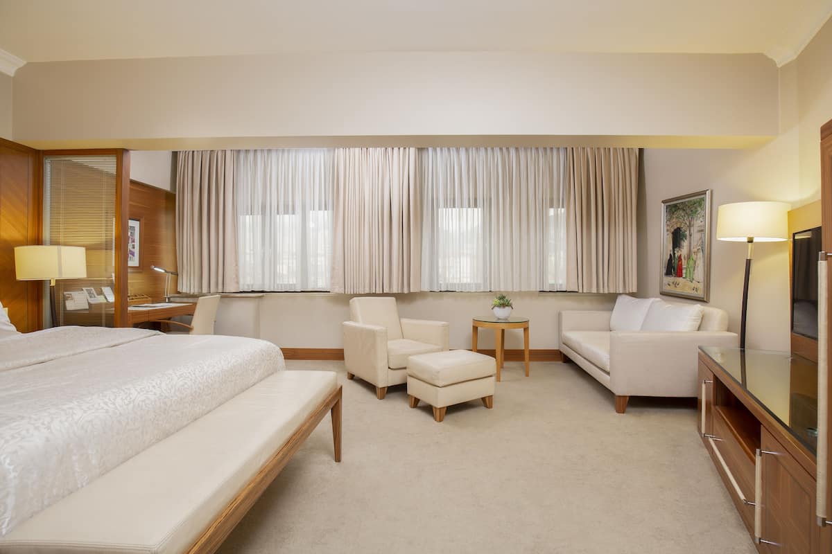 Een hotelkamer met een groot bed, een zithoek met een bank en fauteuils, een tafeltje met een lamp en een televisie.