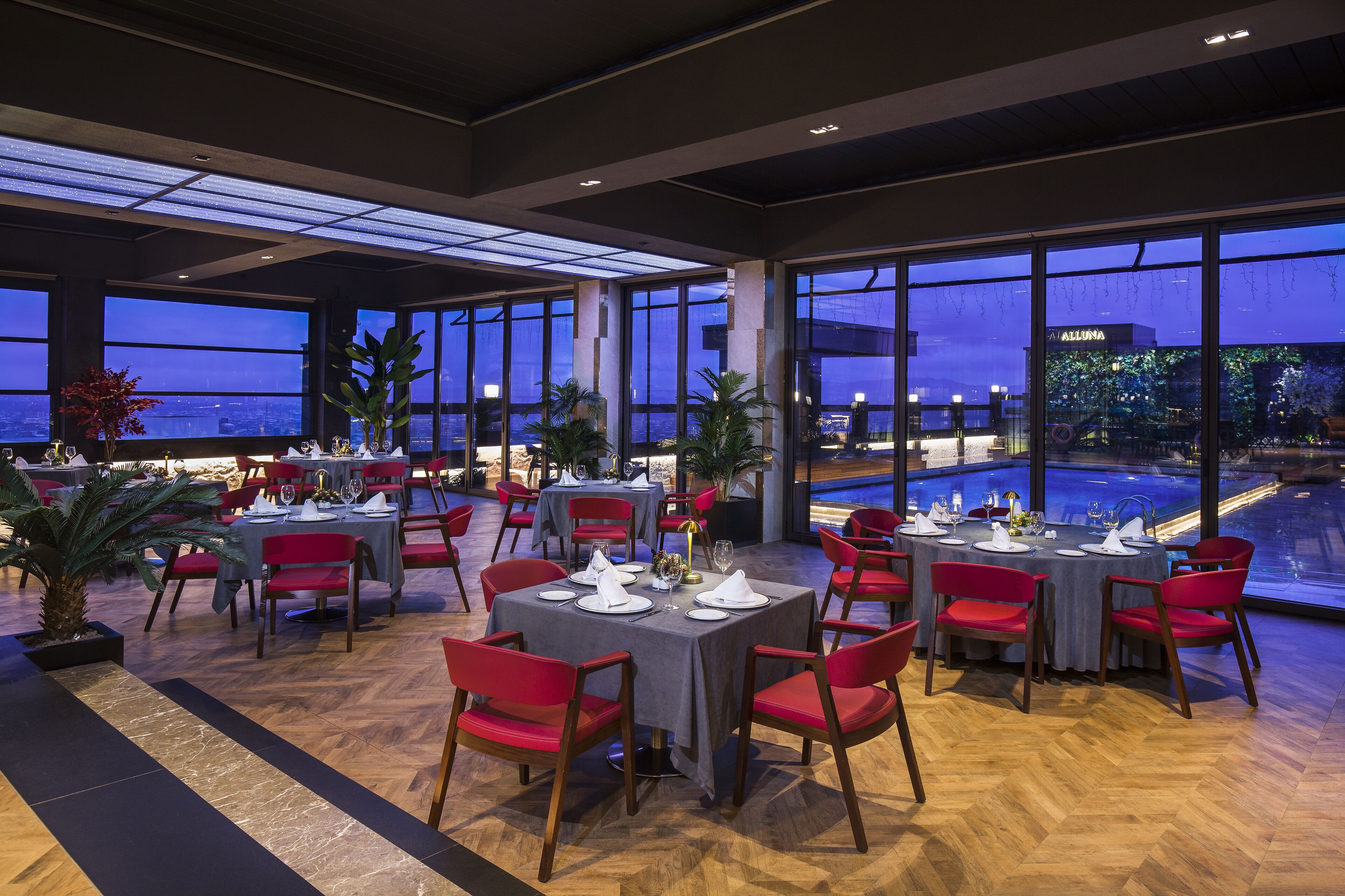 Un restaurante con varias mesas dispuestas para comer, amplias ventanas que ofrecen vistas del paisaje urbano y una zona de piscina visible a través de las ventanas.