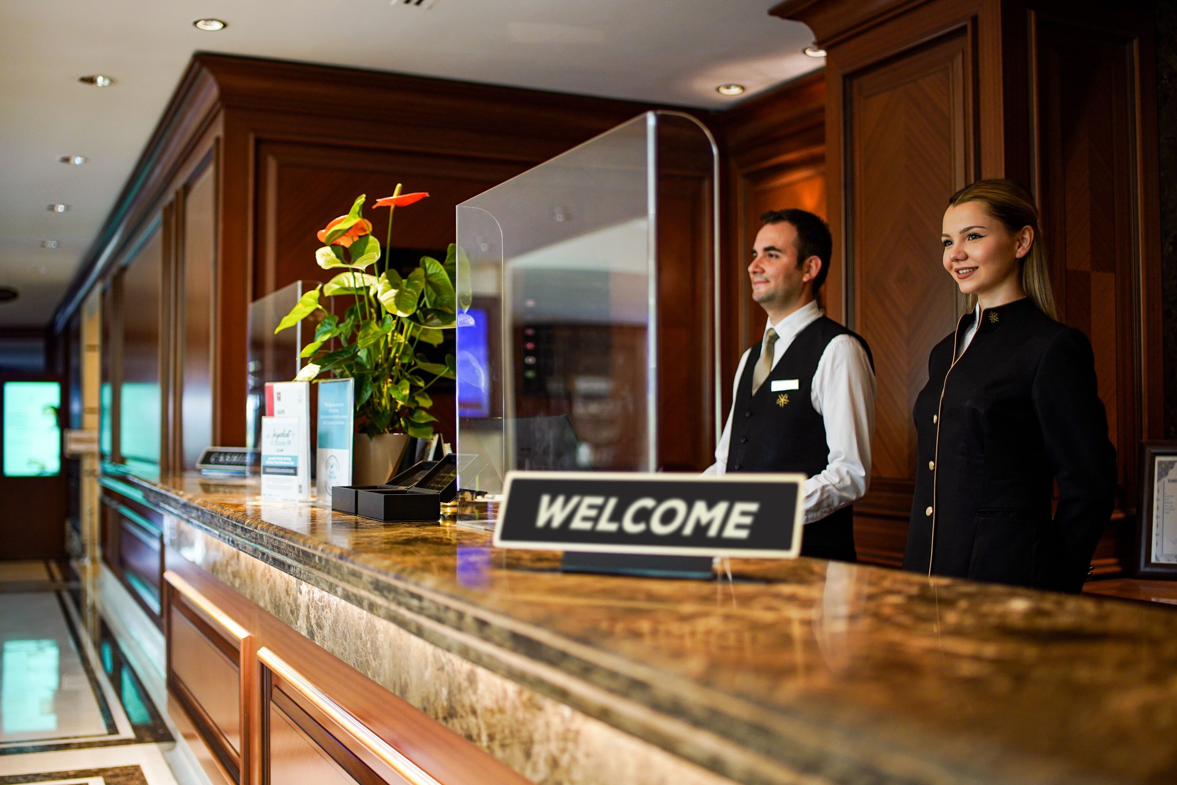A hotel reception desk with a marble counter, a welcome sign, and a potted plant.