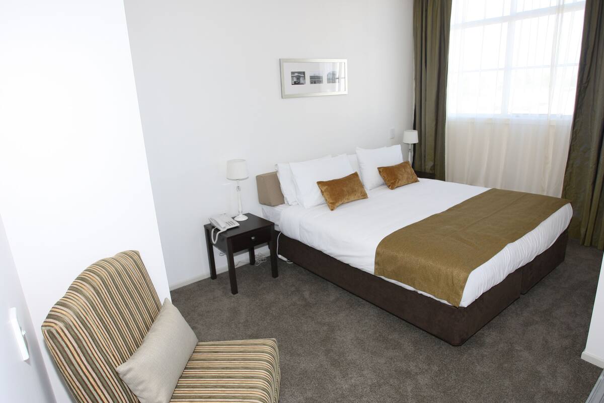 Una habitación de hotel con una cama grande, una mesita de noche con una lámpara, una silla y un cuadro en la pared.