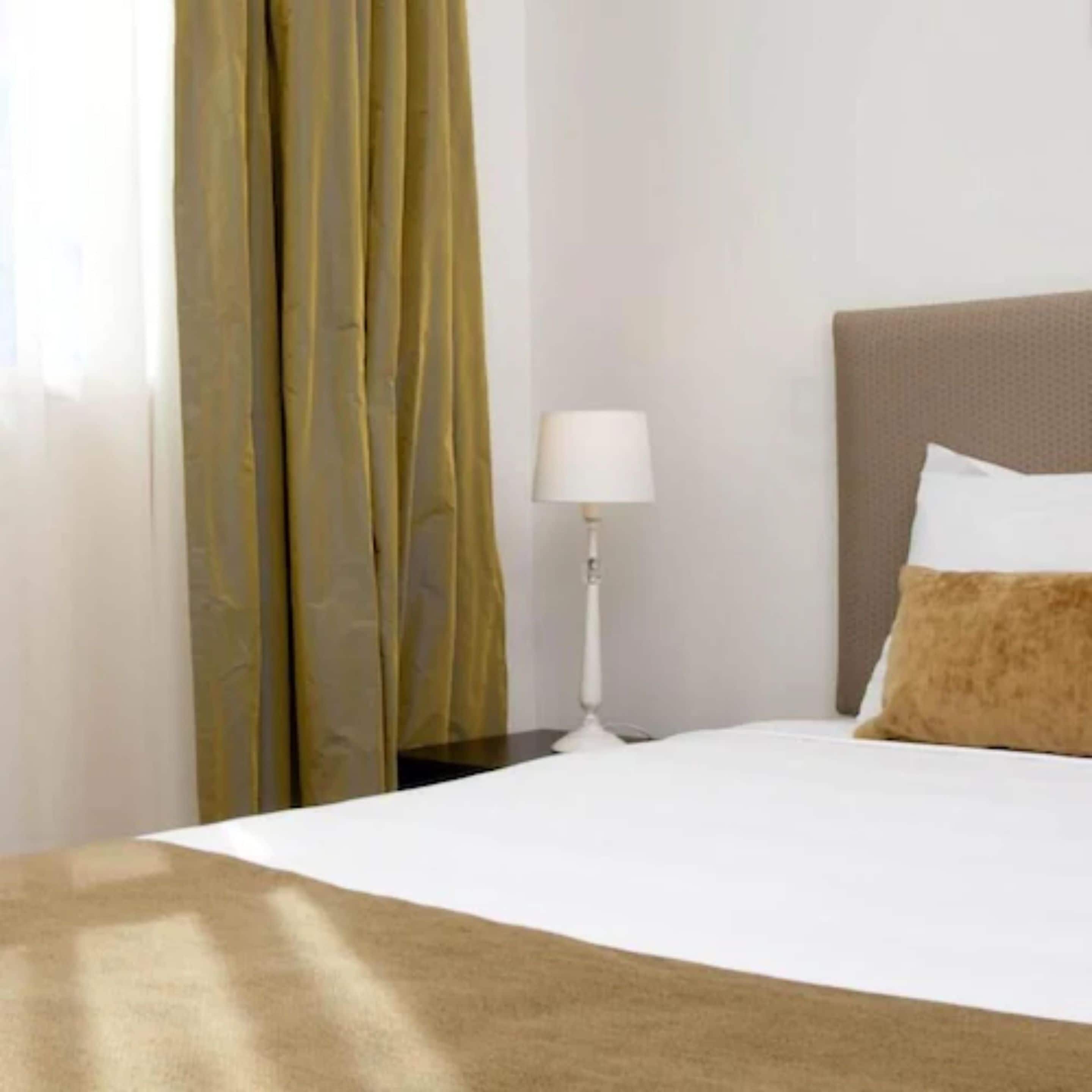 Una camera d'albergo con un letto, un comodino, una lampada e una finestra con tende.