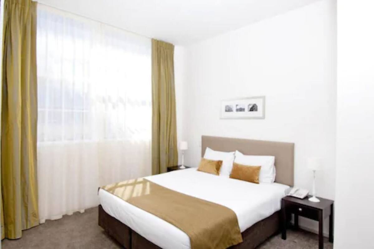 Habitación de hotel con cama, mesita de noche, lámpara y ventana con cortinas.