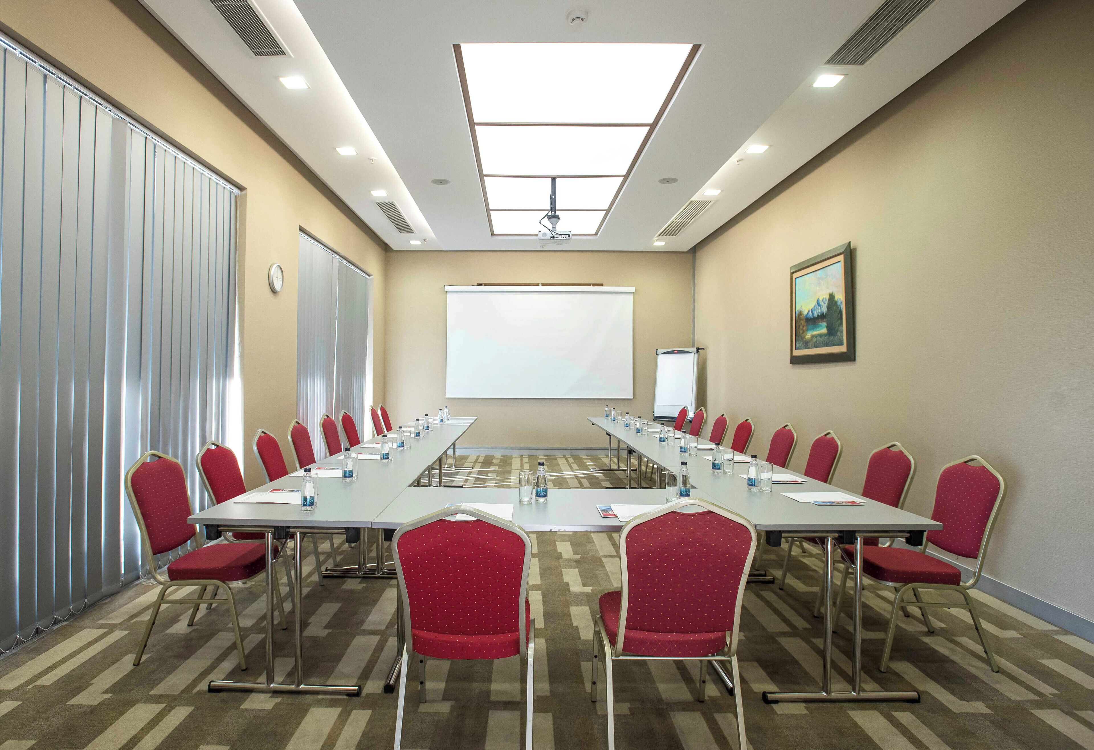 A conference room with a long table, red chairs, a projector screen, and a painting on the wall.