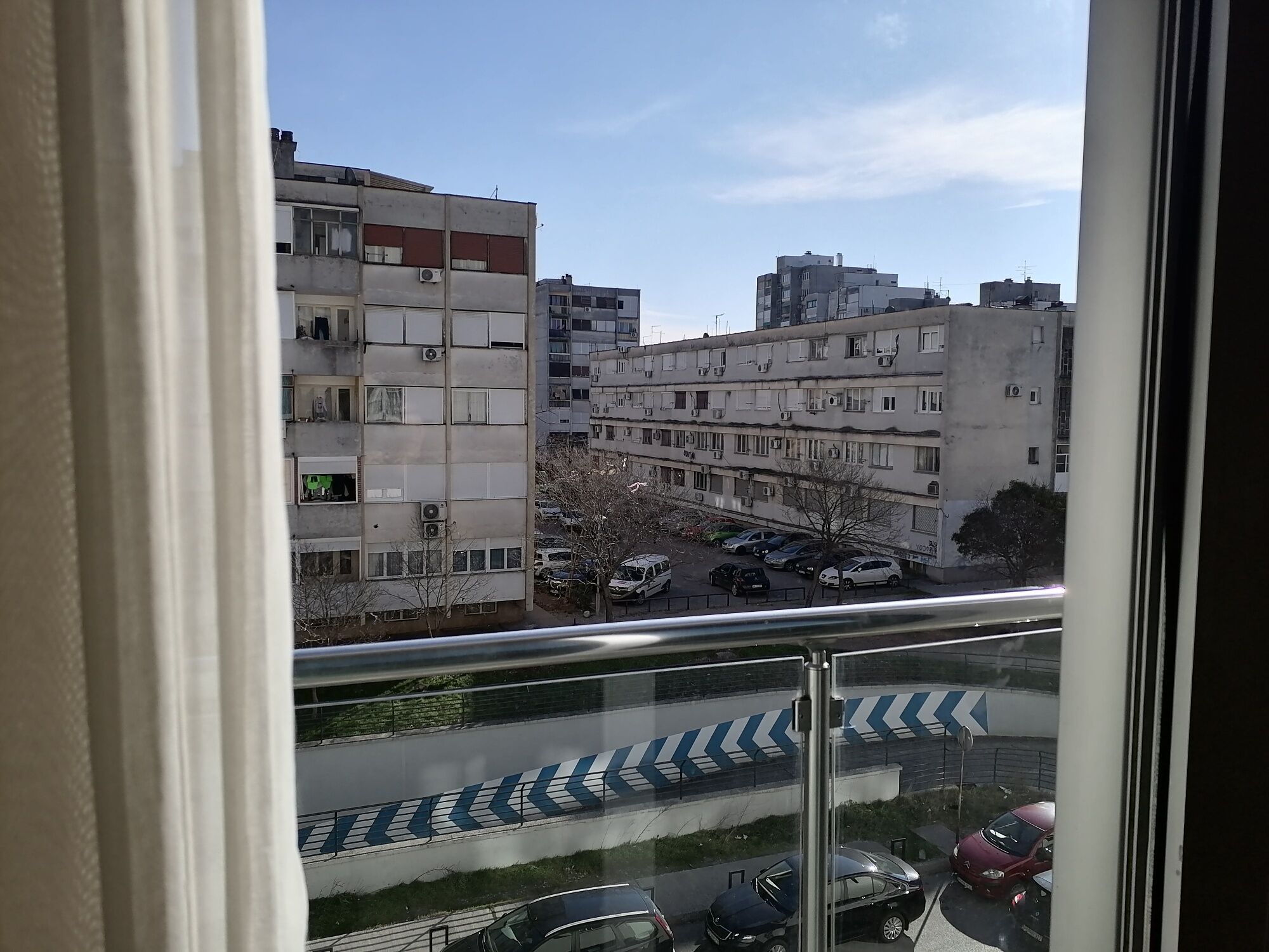 Une vue depuis une fenêtre donnant sur un parking et des immeubles à plusieurs étages, dotés de balcons et de climatiseurs.