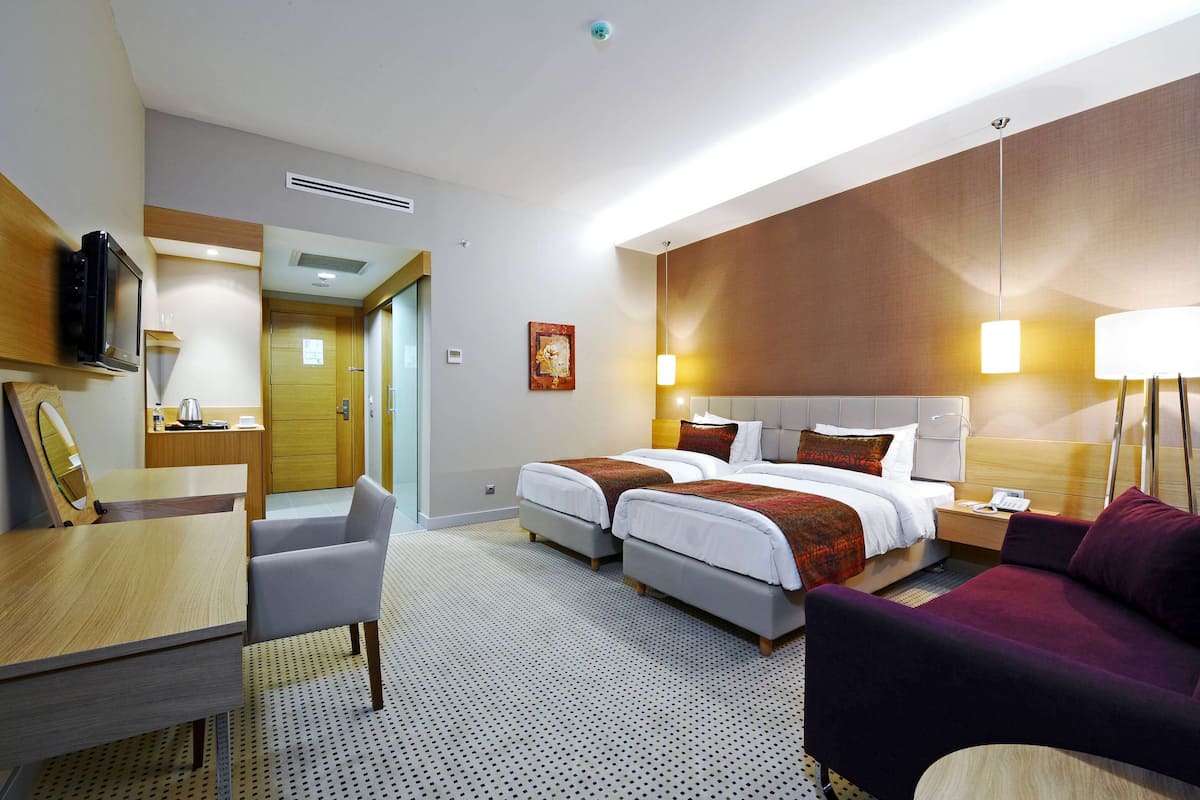 A hotel room with two beds, a desk with a chair, a sofa, and a TV mounted on the wall.