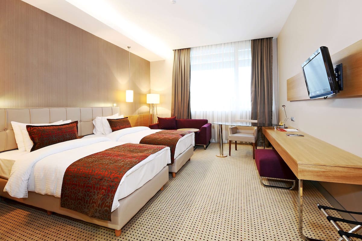 A hotel room with two beds, a desk, a chair, a TV, and a window with curtains.