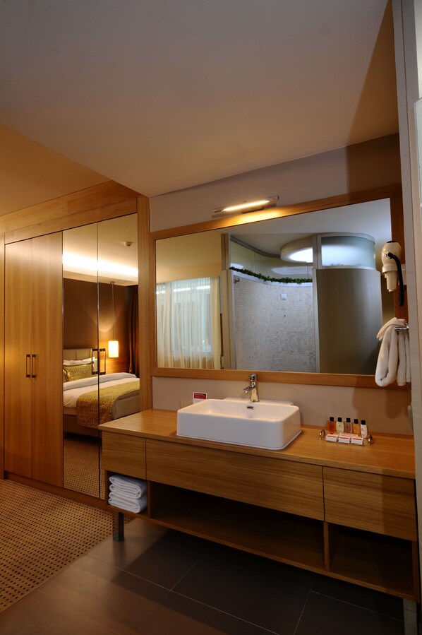 Une salle de bain moderne dans un hôtel, dotée d’un grand miroir, d’un meuble-lavabo en bois et d’une chambre à coucher séparée, visible à travers une porte en verre.