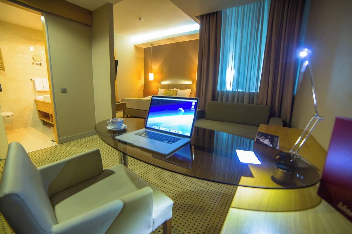 A hotel room with a laptop on a glass table, a sofa, a bed with a headboard, a bedside table with a lamp, and a view of the bathroom.