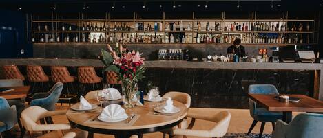 Un restaurante con zona de bar, mesas dispuestas con manteles blancos y sillas, y una variedad de botellas en las estanterías del bar.