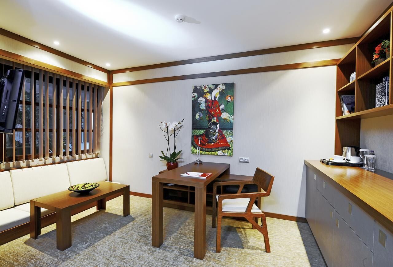A modern dining area with a wooden table, chairs, and a bench. There is a painting on the wall, a potted plant, and a kitchen counter with cabinets.