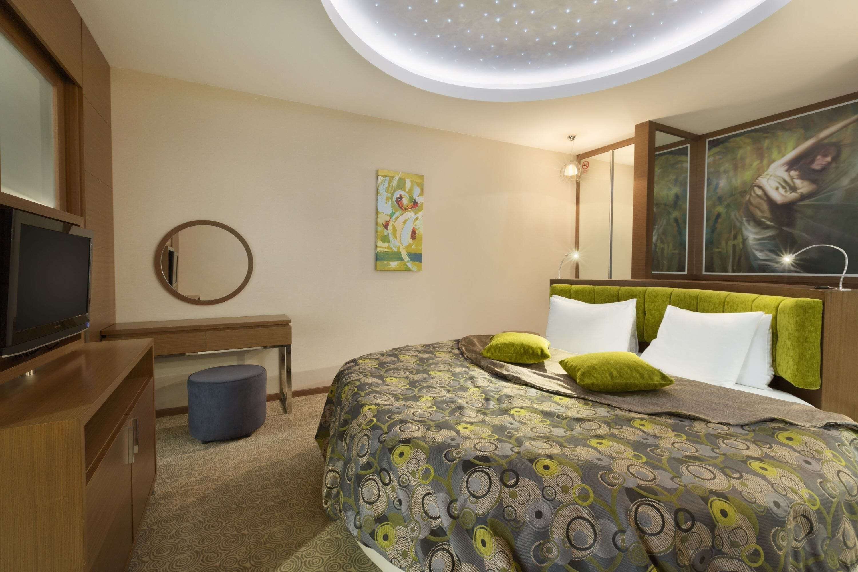 A modern hotel room with a large bed, a television, a desk, and a ceiling light fixture.