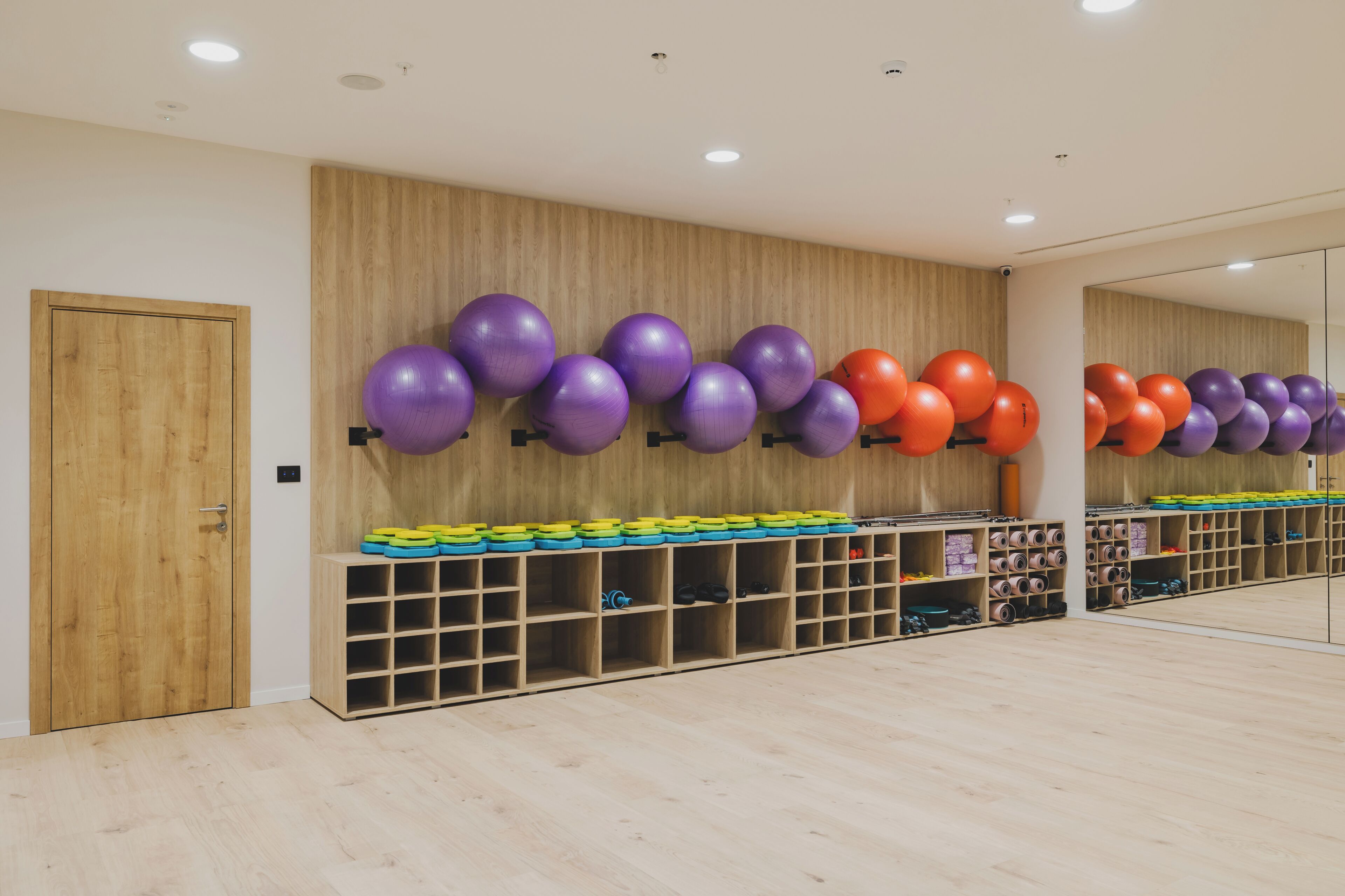 A room with wooden flooring, a mirrored wall, and exercise equipment including purple and orange balls and colorful mats.
