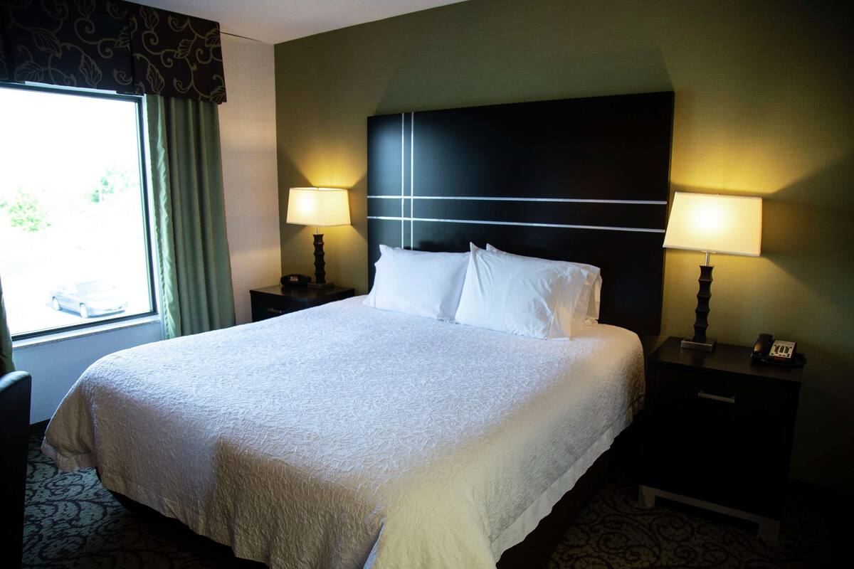 Quarto de hotel com uma cama grande, duas mesas de cabeceira com abajures, uma janela com cortinas e um tapete estampado.