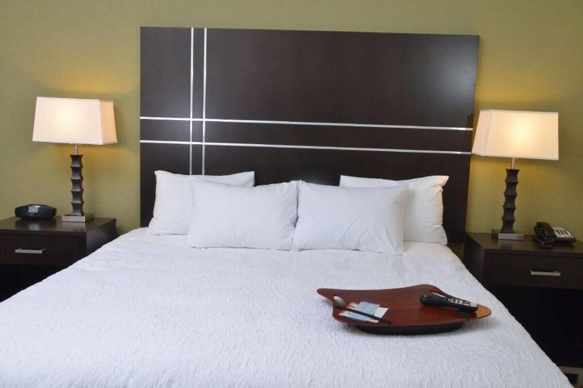 Quarto de hotel com uma cama grande, duas mesas de cabeceira com luminárias, um apoio para controles remotos e uma cabeceira moderna.