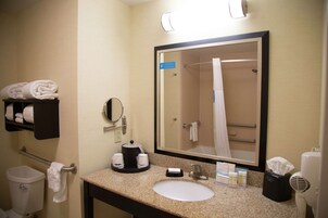 Un baño de hotel con lavamanos, espejo, toallero y una ducha visible en el reflejo del espejo.