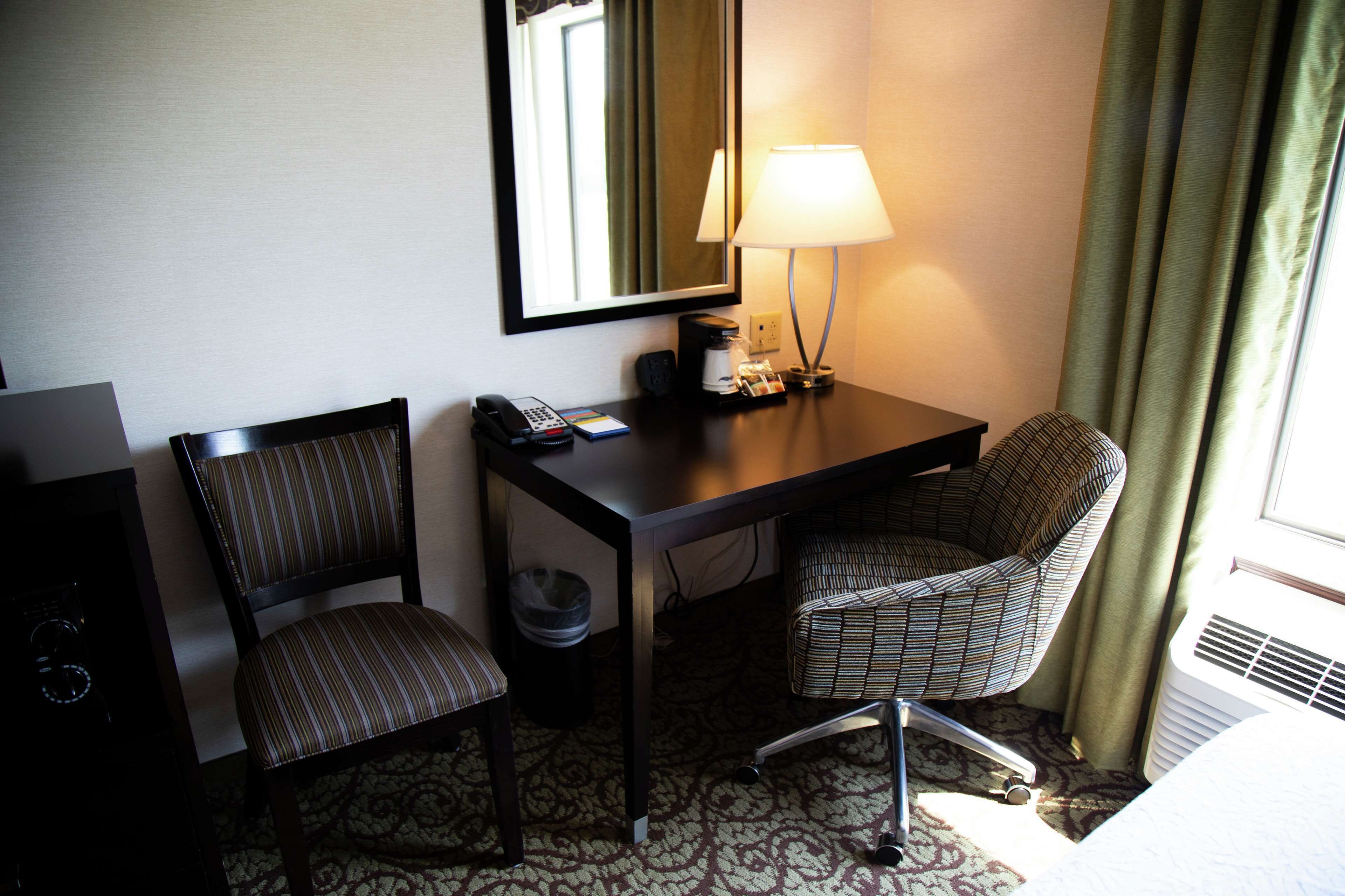 Quarto de hotel com escrivaninha, duas cadeiras listradas, luminária, telefone e lixeira.