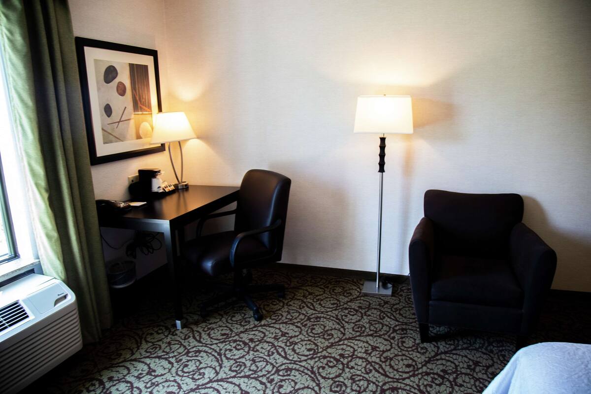 Quarto de hotel com uma secretária, duas cadeiras, um candeeiro de pé e um tapete estampado.