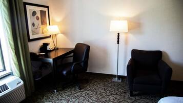 Una habitación de hotel con escritorio, dos sillas, una lámpara de pie y una alfombra estampada.