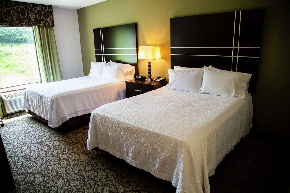 Quarto de hotel com duas camas, uma mesa de cabeceira com abajur e vista para o verde lá fora.