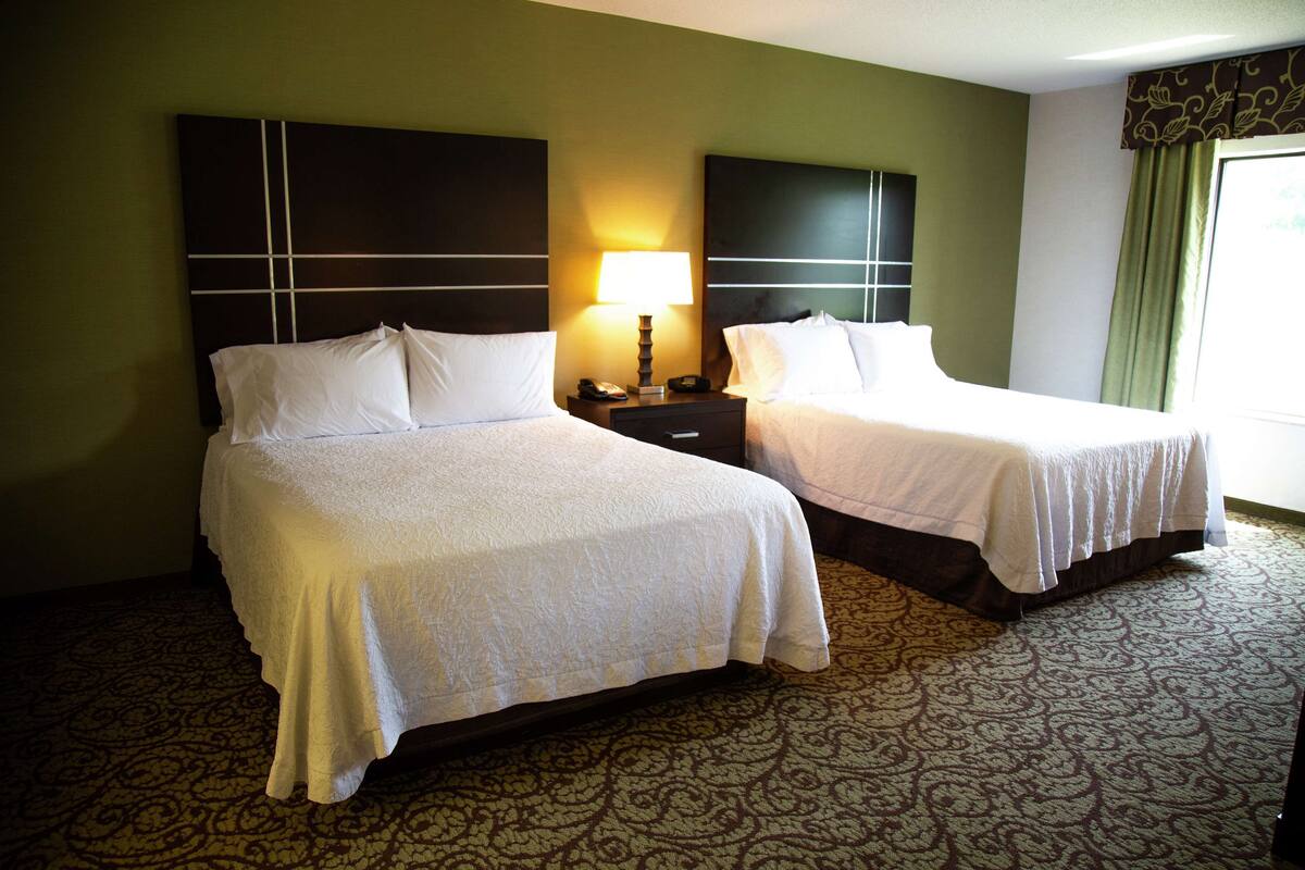 Quarto de hotel com duas camas, uma mesa de cabeceira com abajur e um tapete estampado.