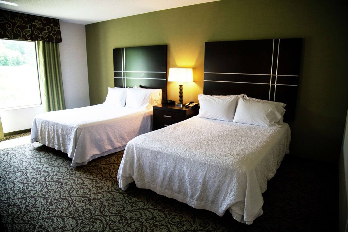 Quarto de hotel com duas camas, uma mesinha de cabeceira com abajur e um tapete estampado.