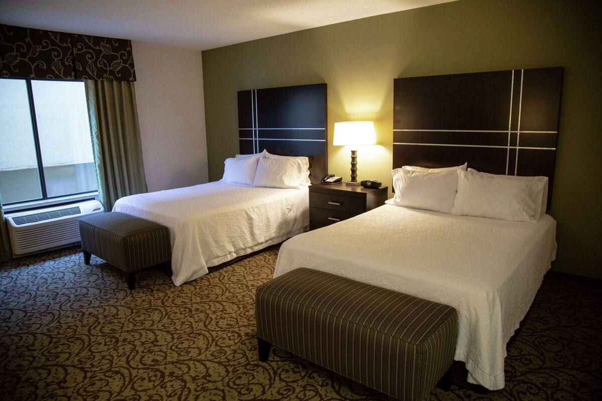 Quarto de hotel com duas camas, janela com cortinas, mesa de cabeceira com abajur e carpete estampado.
