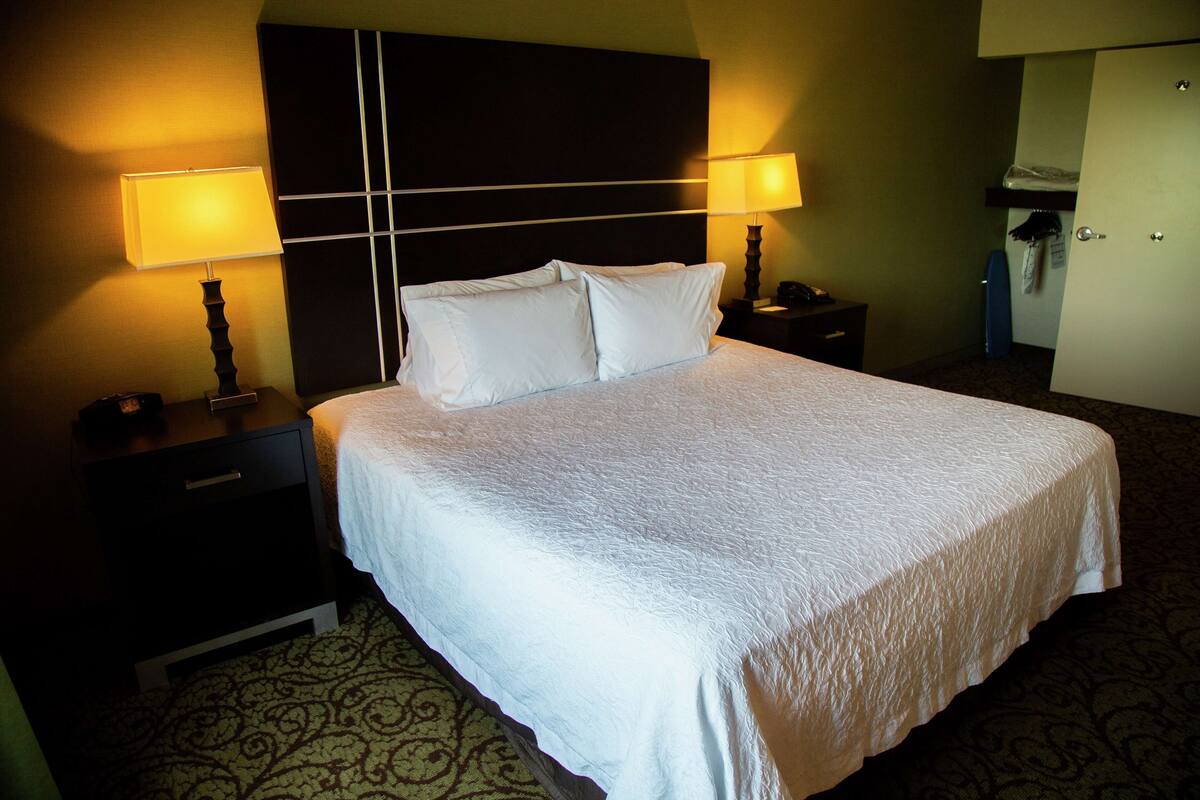 Quarto de hotel com uma cama grande, duas mesas de cabeceira com luminárias, um guarda-roupa e um tapete estampado.